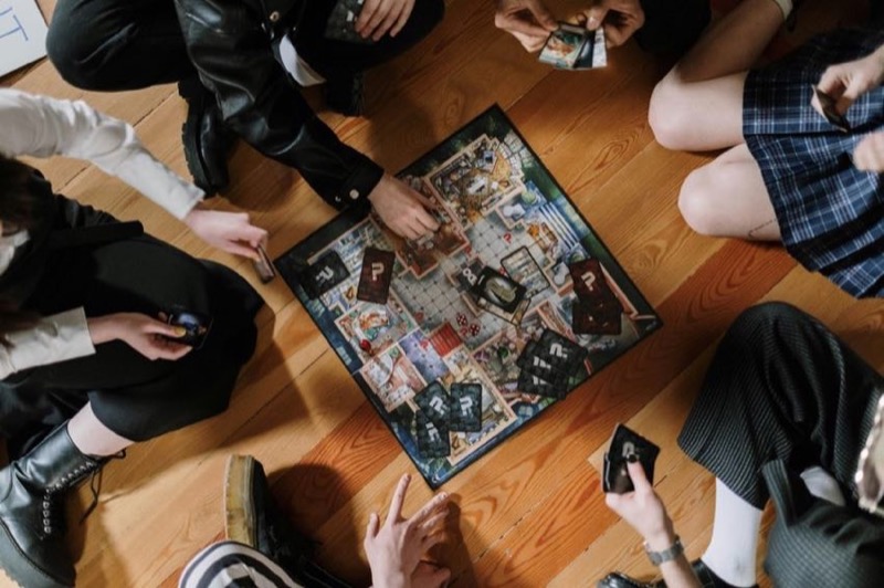Overhead view of six friends sitting on a wood floor around a mystery board game with cards spread out