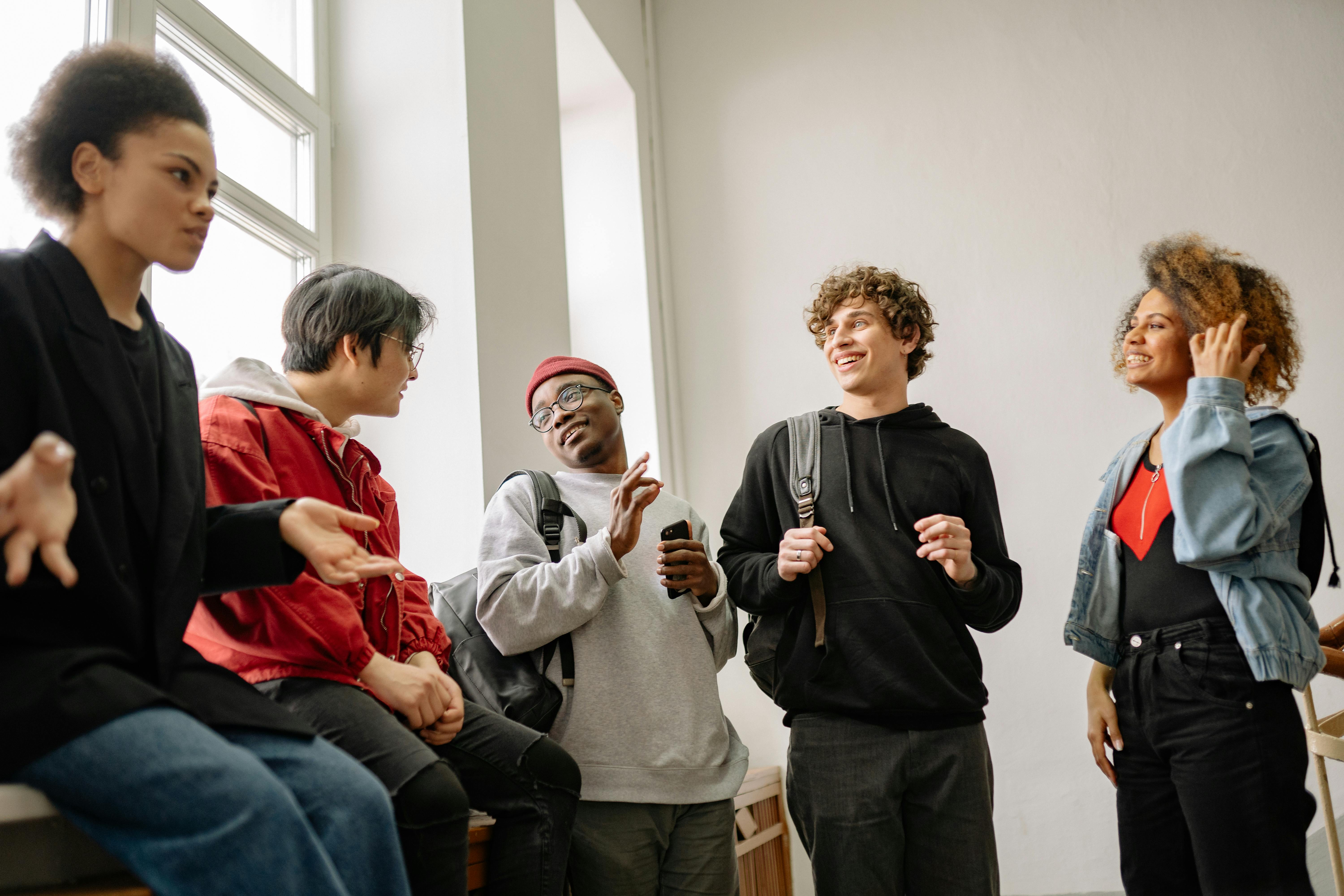 Five diverse friends talking and laughing together indoors by a large window, representing genuine connection