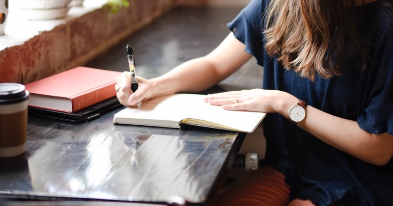A young woman journaling at a sunlit table with coffee and a book beside her
