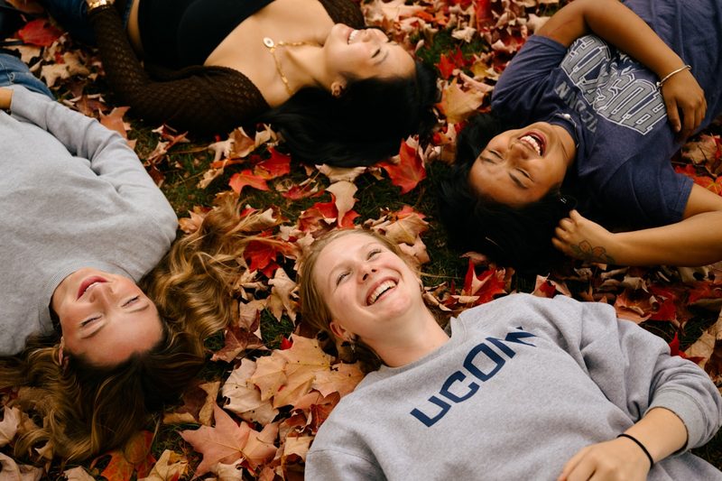 Four women laughing together outdoors surrounded by autumn leaves