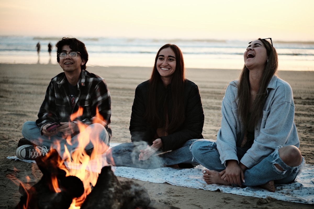 Three friends sitting around a beach fire at sunset, laughing together