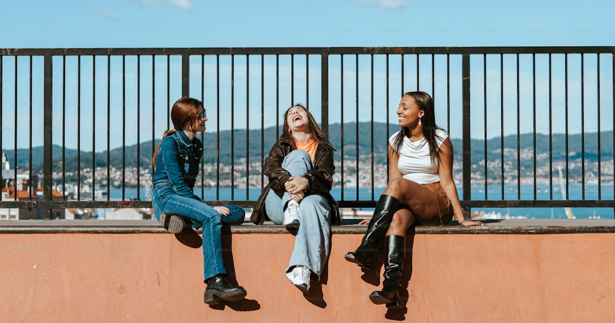Three women sitting on a wall or ledge with a city and bay behind them, laughing in the sunshine