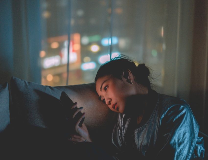 Woman scrolling her phone alone on the couch at night with a blurred city skyline glowing through the window behind her