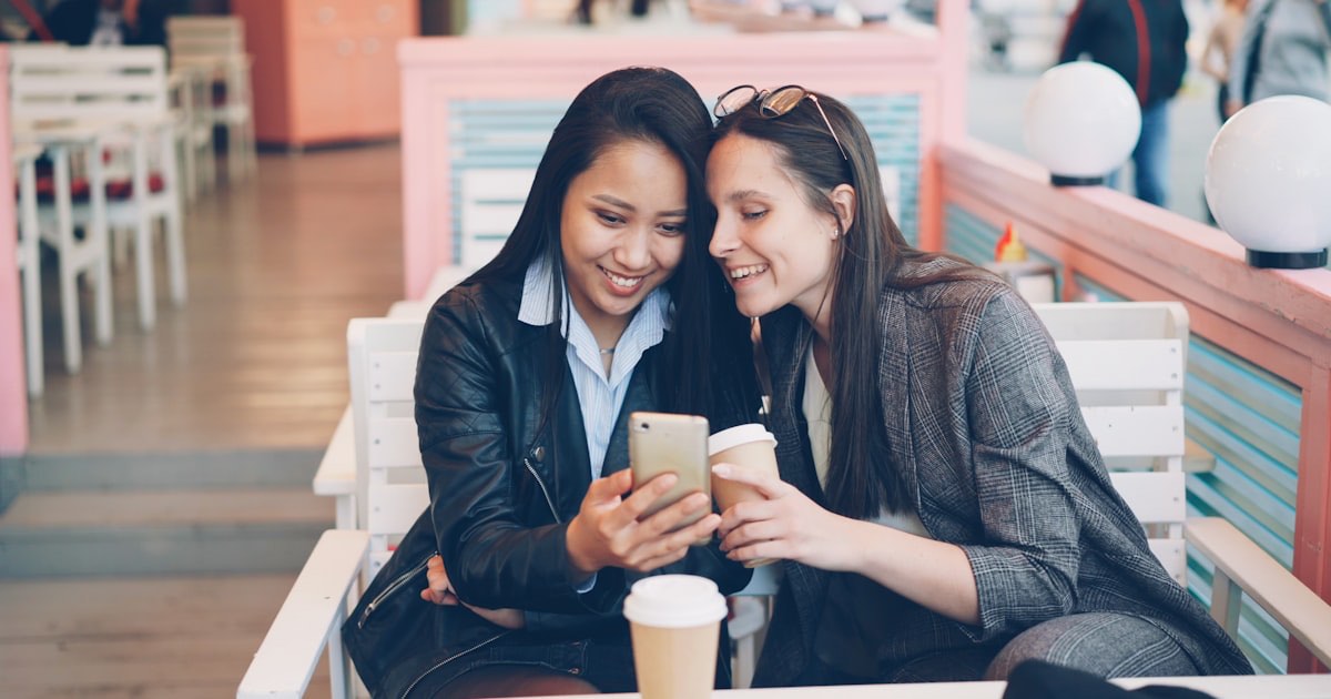 Two friends leaning in close over a phone at an outdoor cafe table, both smiling, the kind of screen-sharing moment that turns a shared fandom into an actual conversation