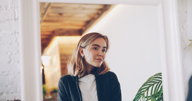 A young woman with shoulder-length blonde hair, seen in a white-framed mirror at home, smiling slightly beside a leafy plant.